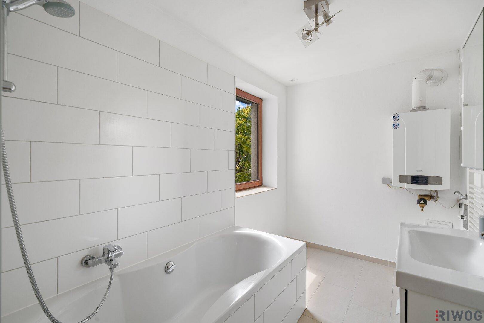 Helles Badezimmer mit Badewanne und Duscharmatur, weißen Fliesen und Fenster.