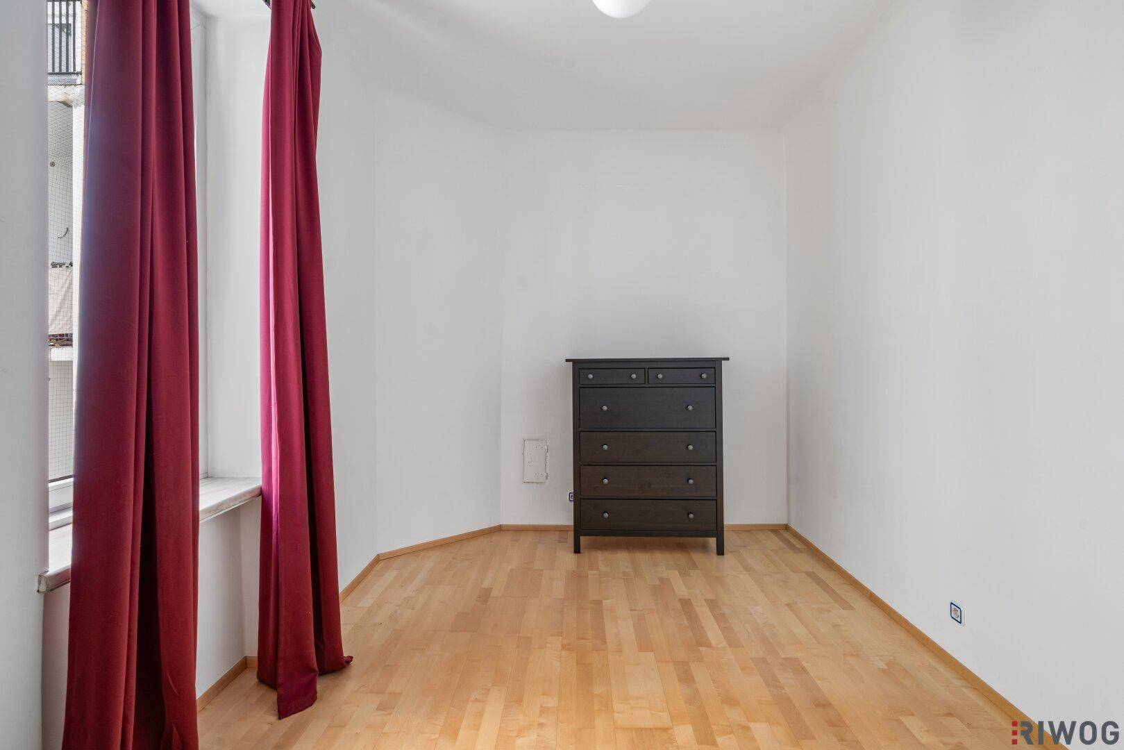 Schlafzimmer mit Holzboden, einem Fenster mit roten Vorhängen und einer Kommode.