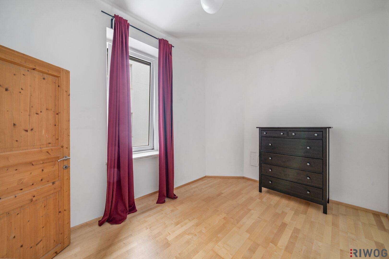 Helles Schlafzimmer mit Holzboden, einer Holztür und einem Fenster mit roten Vorhängen.