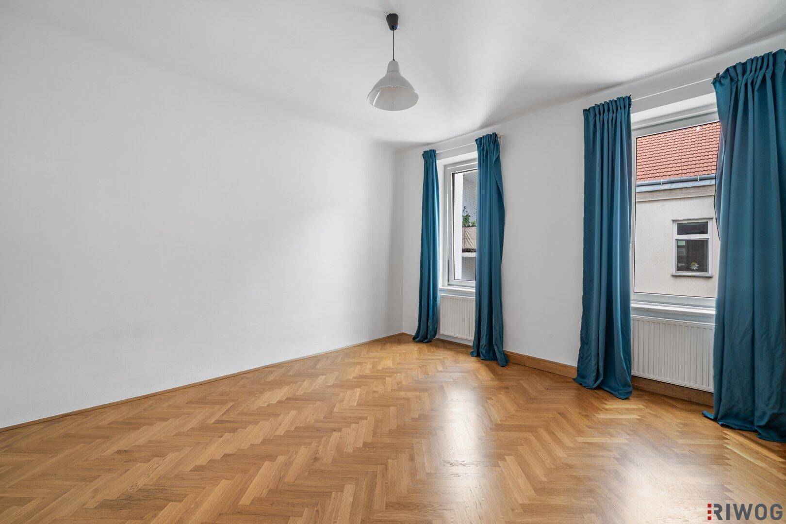 Geräumiges Wohnzimmer mit Fischgrätparkett, zwei Fenstern und blauen Vorhängen.