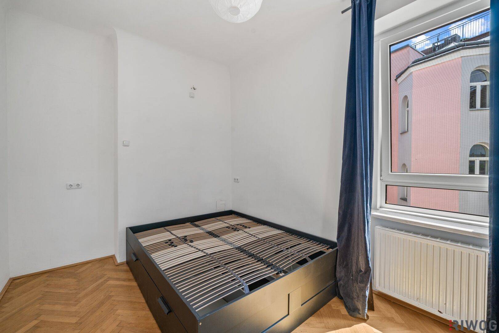 Schlafzimmer mit Fischgrätparkett und einem Bettgestell mit Stauraum unter dem Fenster.