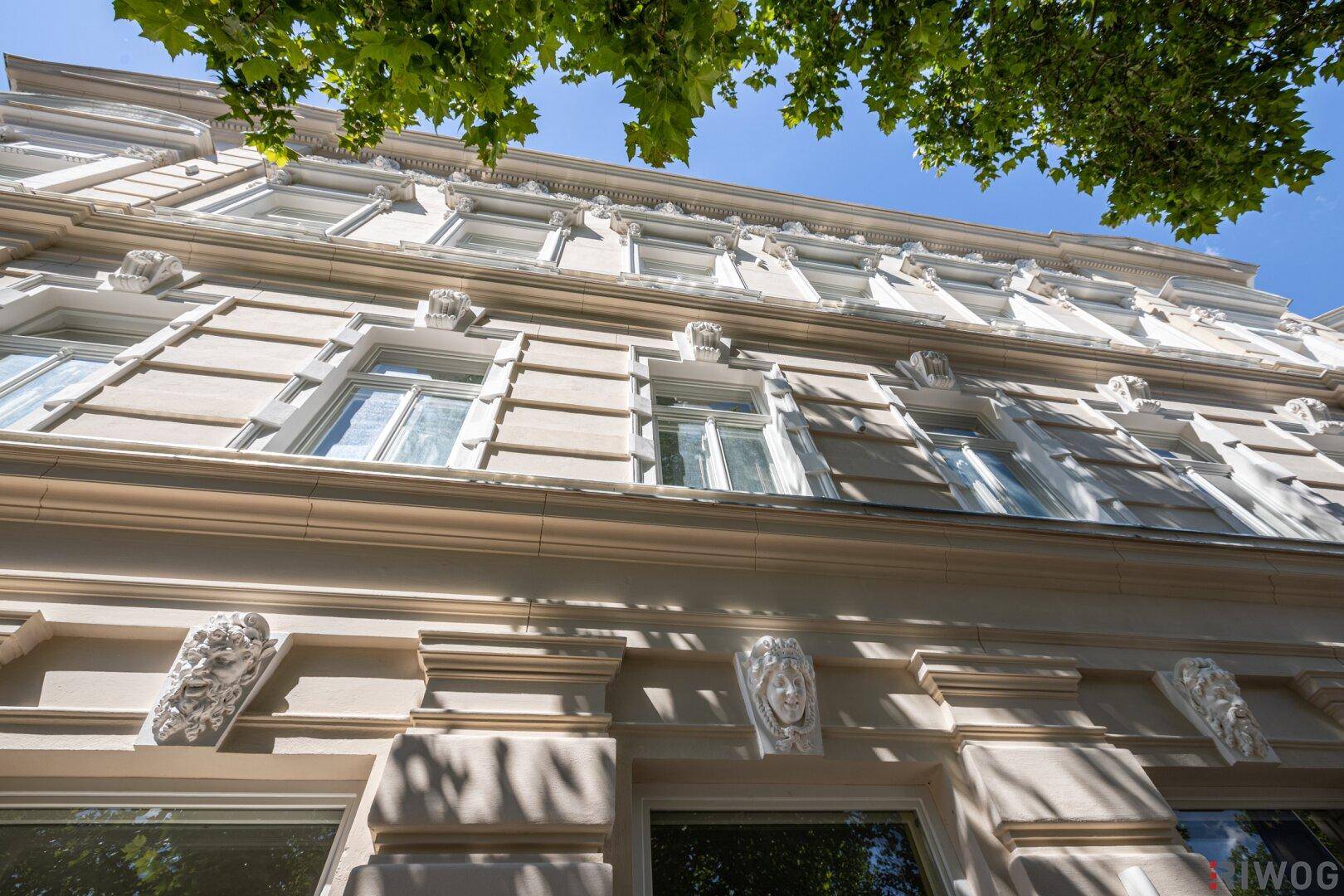 Detailansicht der historischen Fassade mit aufwendigen Stuckverzierungen und großen Fenstern unter blauem Himmel.