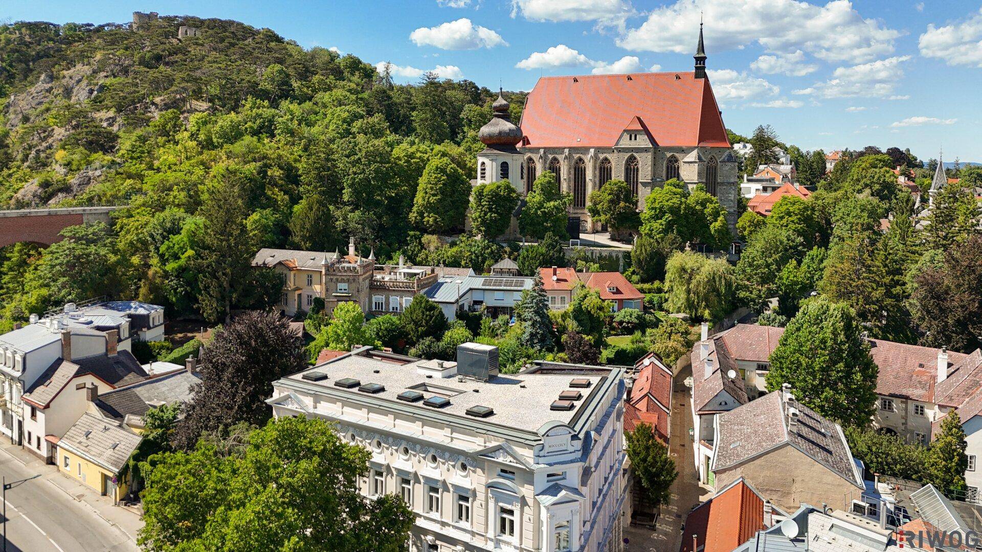 Weitläufige Luftaufnahme der Stadt bei Tageslicht mit dem Gebäude, einer Kirche und grünen Hügeln.