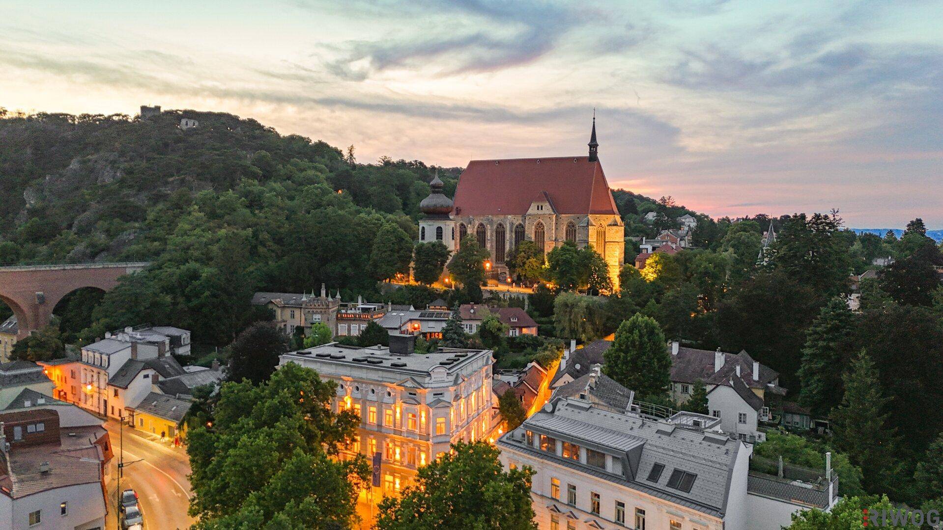 Panoramablick auf eine historische Stadt bei Sonnenuntergang mit beleuchteten Gebäuden und grünen Hügeln.