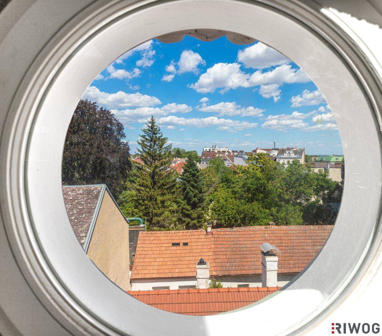 Blick aus einem runden Fenster auf die Dächer der Stadt und den blauen Himmel mit Wolken.