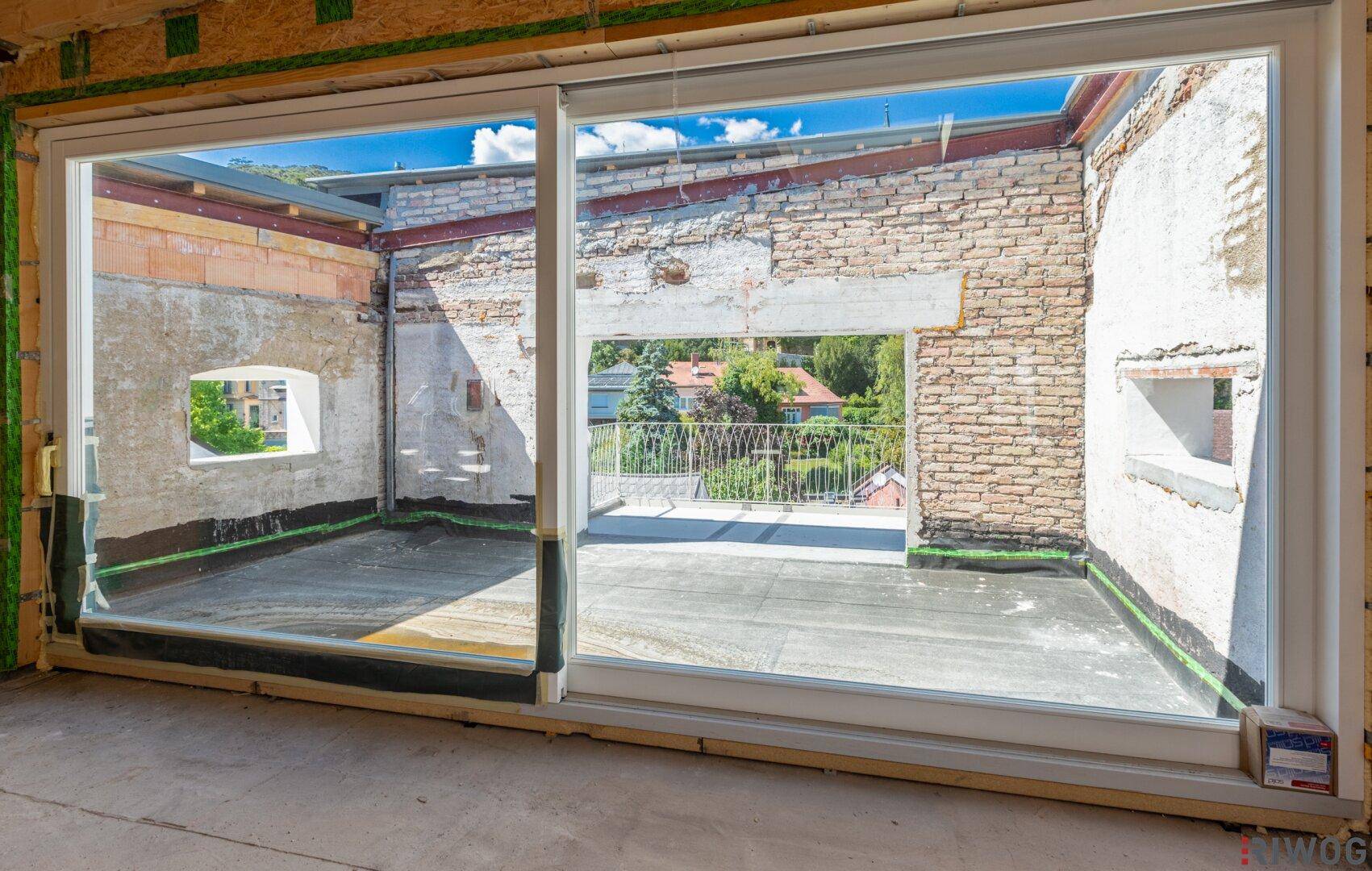 Rohbau eines Raumes mit großer Fensterfront und Zugang zu einem zukünftigen Balkon.
