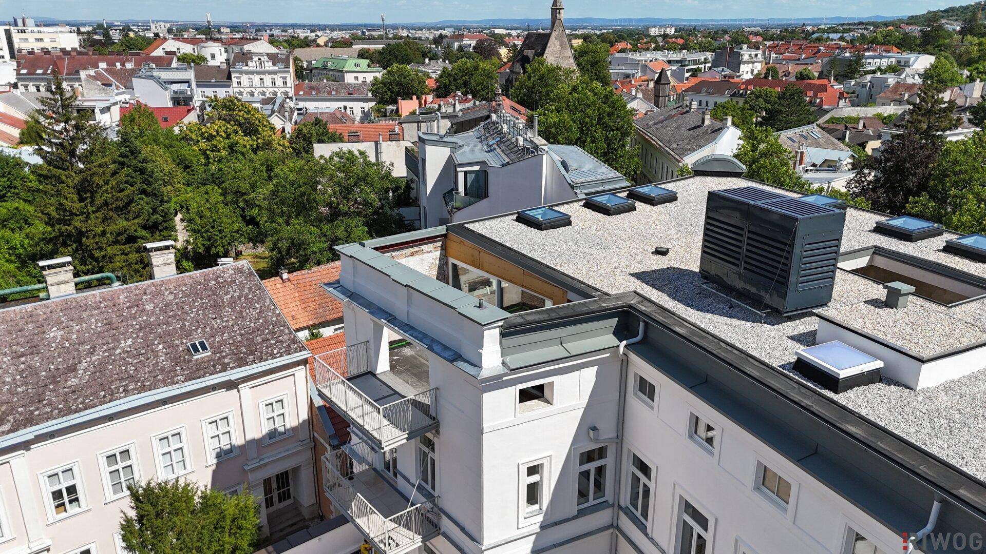 Luftaufnahme eines Gebäudes mit Flachdach und Dachfenstern, umgeben von städtischer Bebauung.