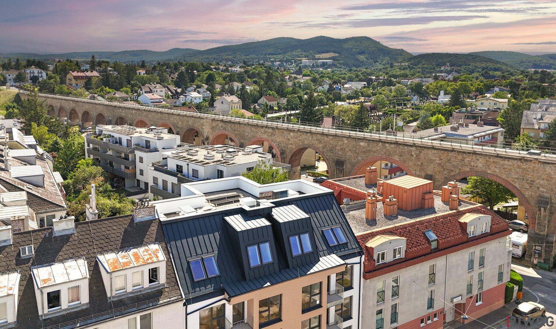 Luftaufnahme des modernen Apartmentgebäudes mit historischem Aquädukt und grüner Hügellandschaft im Hintergrund.