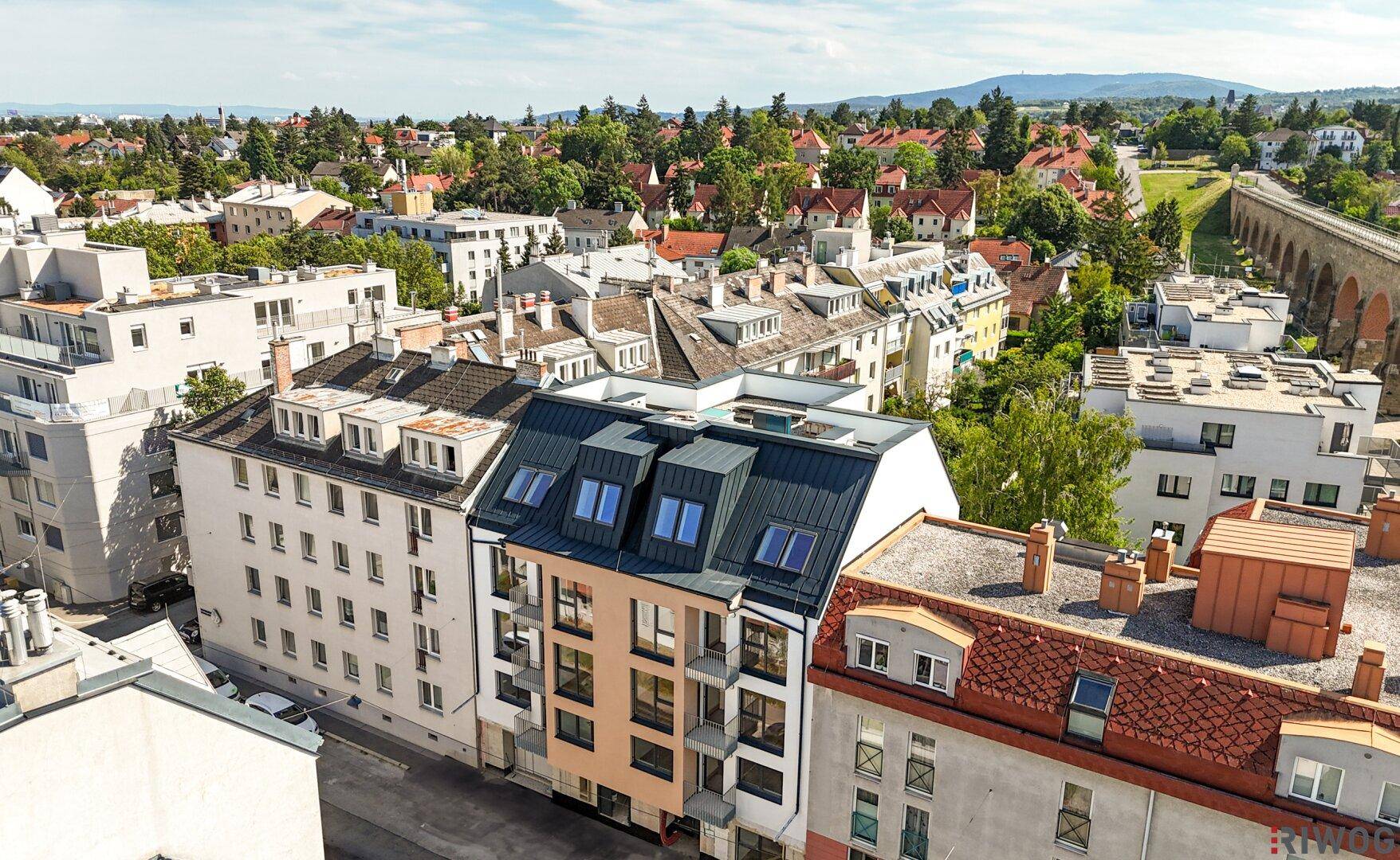 Luftaufnahme des modernen Apartmentgebäudes in städtischer Umgebung mit umliegenden Häusern und viel Grün.