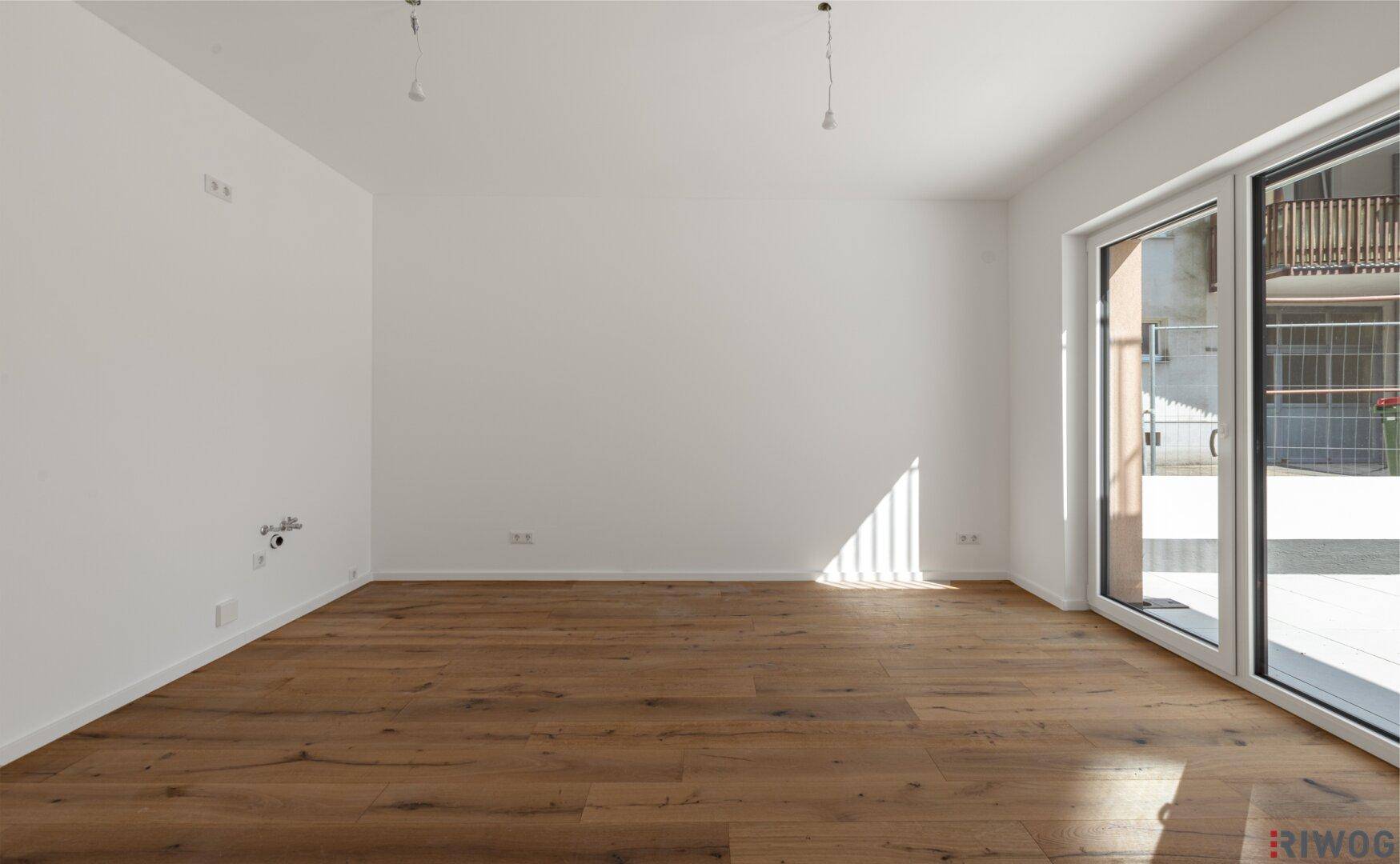 Geräumiges, leeres Wohnzimmer mit hellem Holzboden und großen Fenstern, die viel Tageslicht hereinlassen.