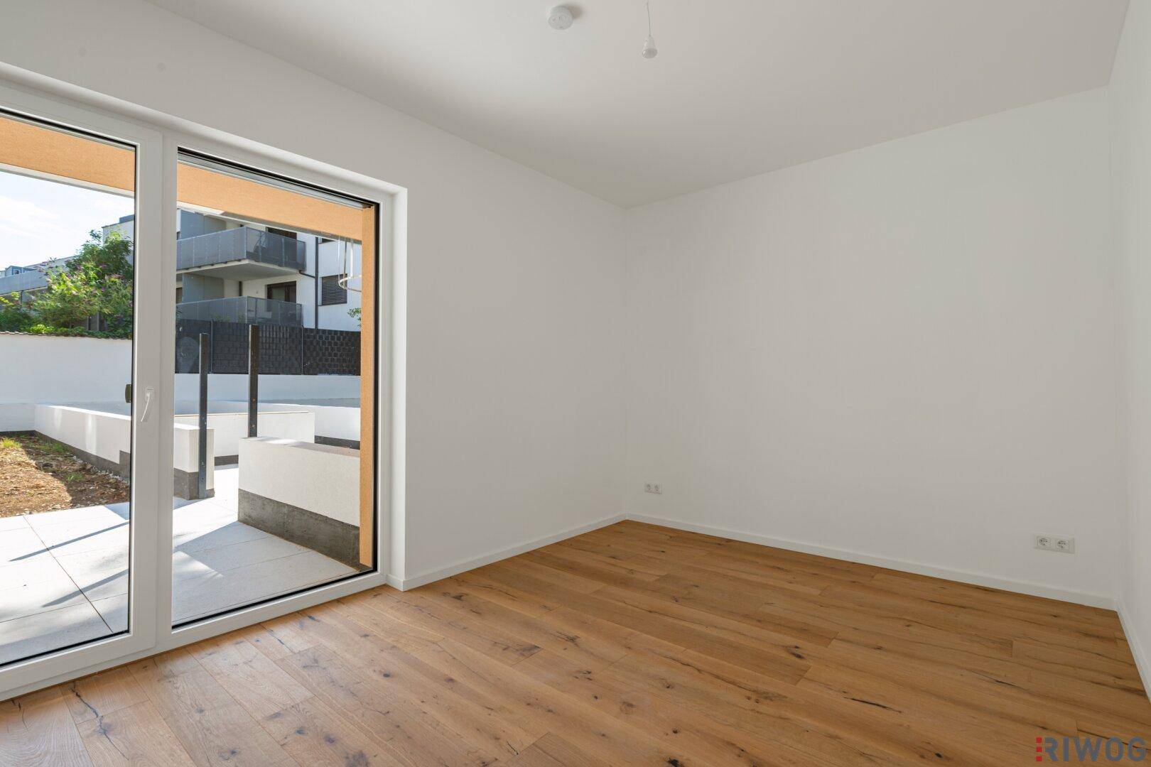 Helles, leeres Schlafzimmer mit Holzboden und großem Fenster, das Zugang zu einem Außenbereich bietet.