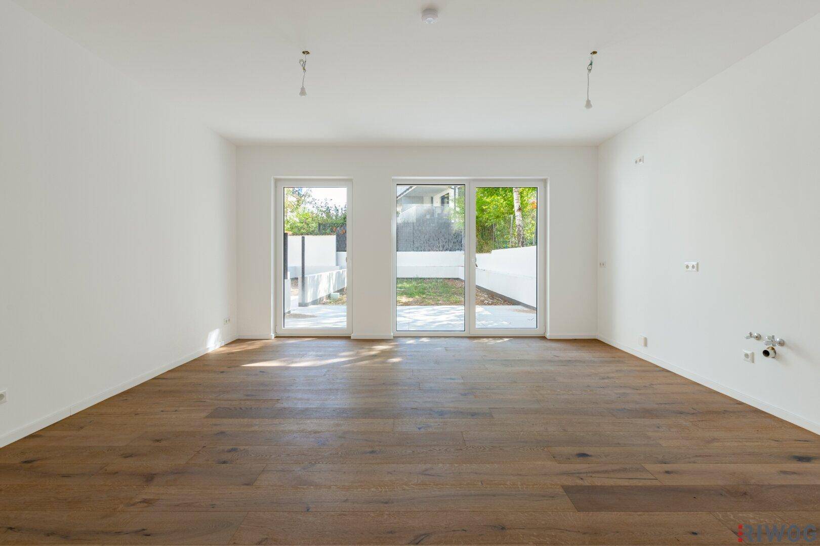 Heller, leerer Wohnraum mit Holzboden und großen Fenstern zum Garten.