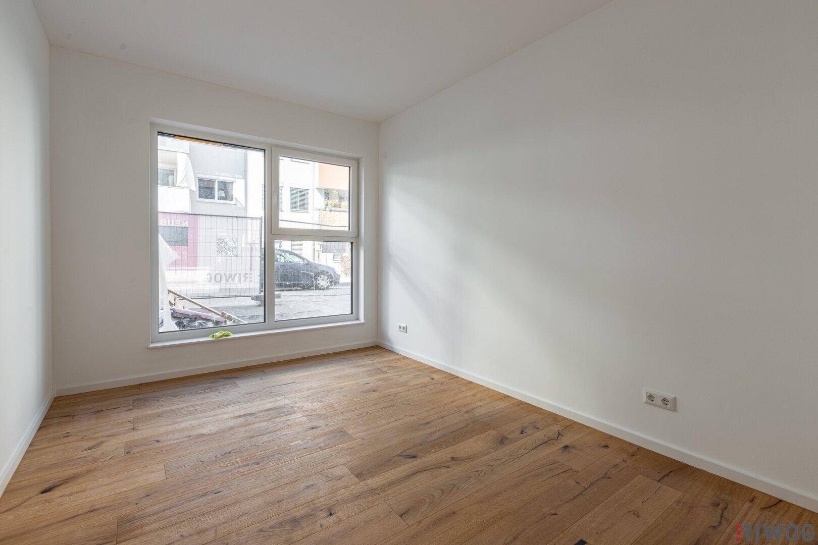Helles, unmöbliertes Zimmer mit Holzboden und großem Fenster zur Straße.