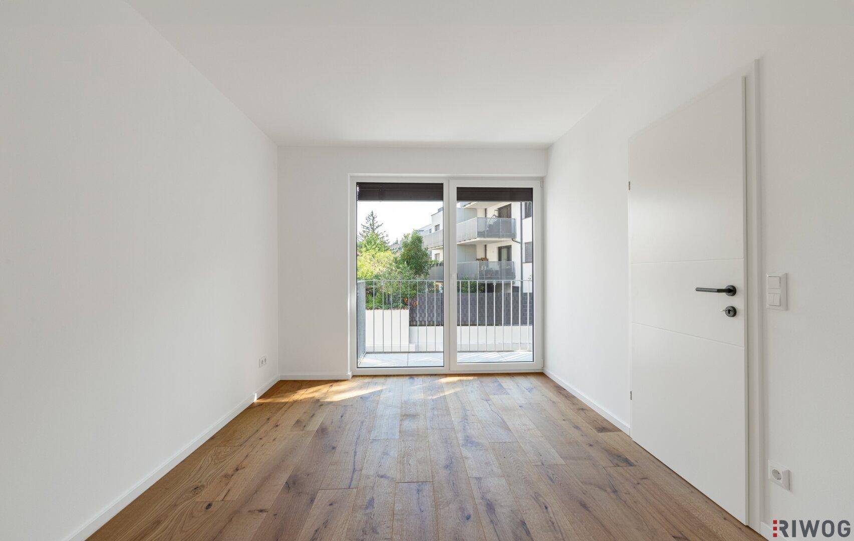 Leeres Schlafzimmer mit Holzboden und großen Fenstern, die Zugang zum Balkon bieten.