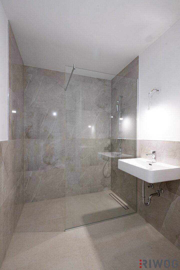 Modernes Badezimmer mit begehbarer Dusche, grauen Fliesen und minimalistischem Waschbecken.