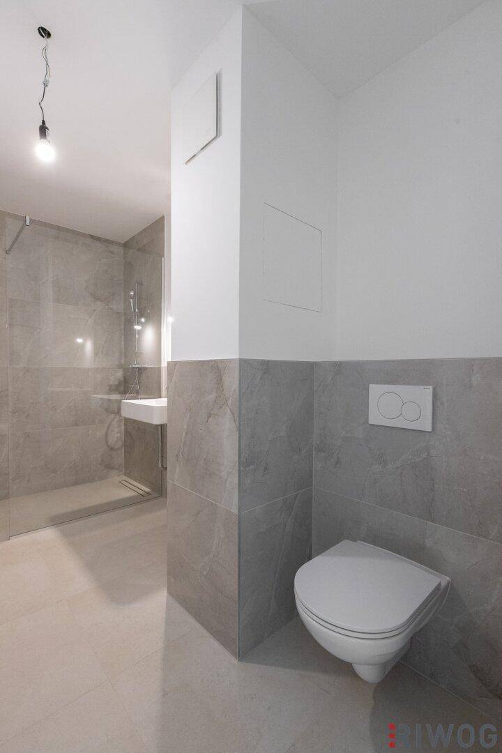 Helles Badezimmer mit wandhängendem WC, grauen Fliesen und begehbarer Dusche im Hintergrund.