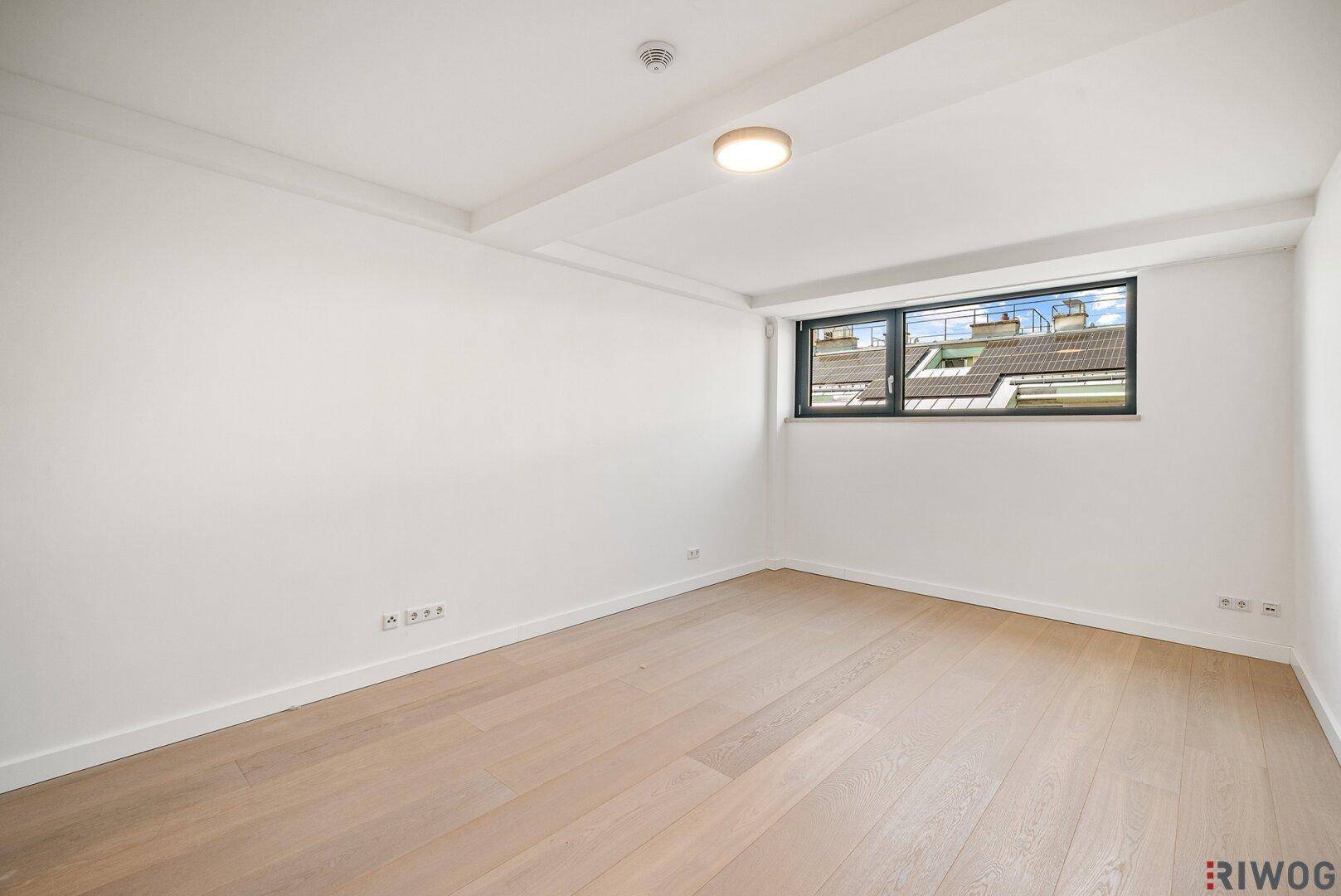 Geräumiges Zimmer mit Holzboden und einem breiten Fenster, das viel Tageslicht hereinlässt.
