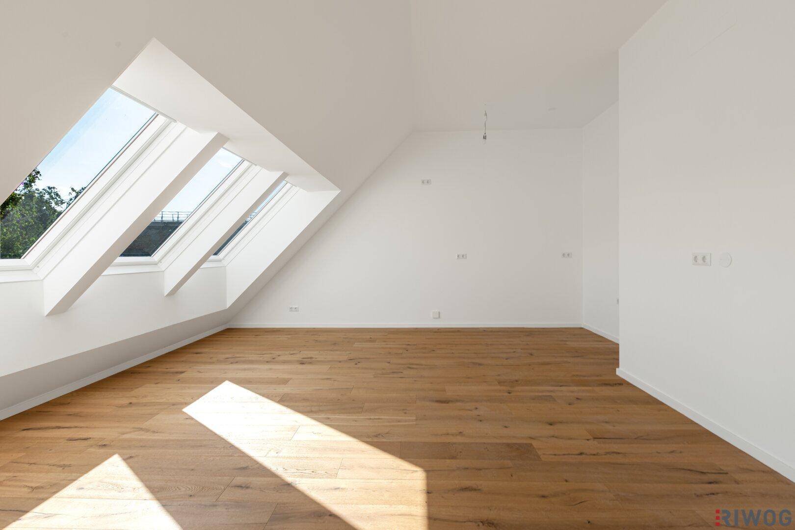 Geräumiger Dachgeschossraum mit schrägen Wänden, Holzboden und viel Tageslicht durch Dachfenster.
