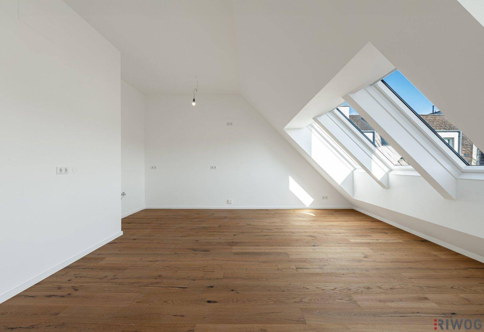 Geräumiger Dachgeschossraum mit Holzboden und schrägen Wänden, durchflutet von natürlichem Licht.