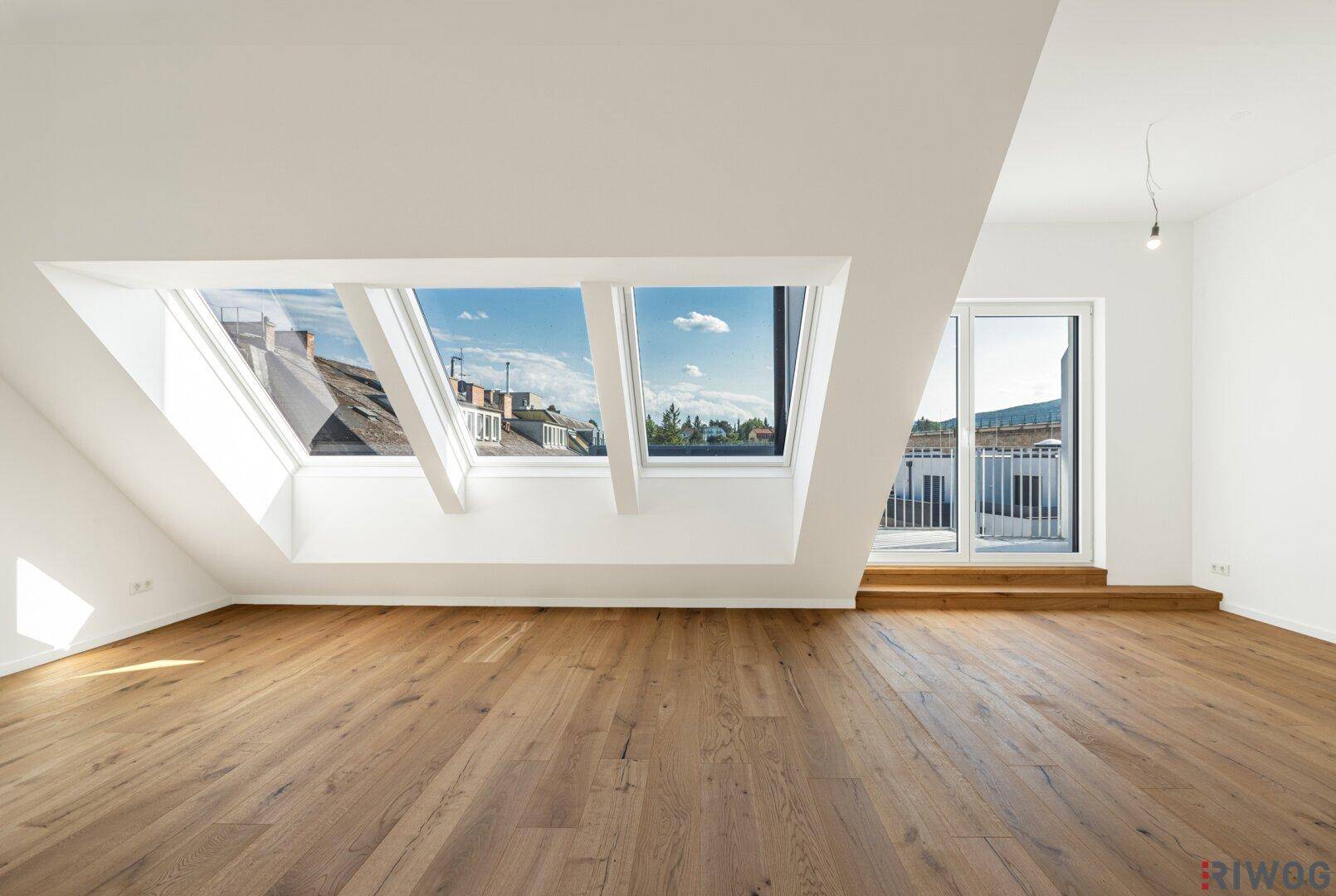 Heller Raum im Dachgeschoss mit Holzboden, großen Dachfenstern und Zugang zu einer Terrasse.