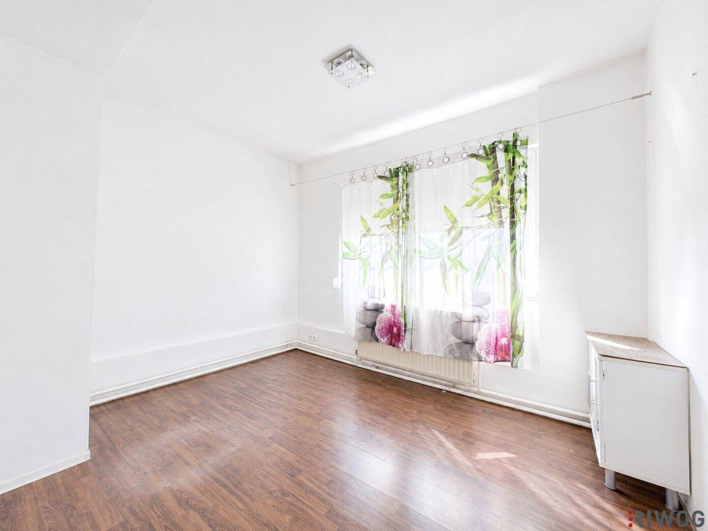 Helles Zimmer mit weißen Wänden, Laminatboden und Fenster mit Vorhängen.