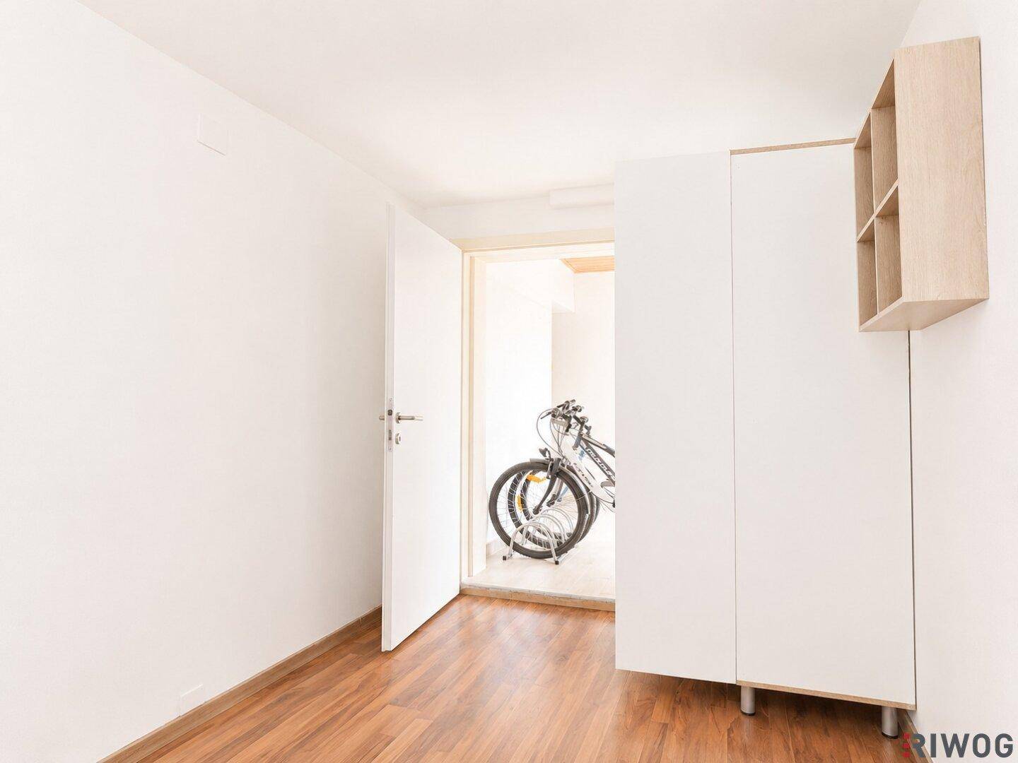 Heller Raum mit Holzboden und Schrank, mit Blick auf einen Fahrradabstellbereich.