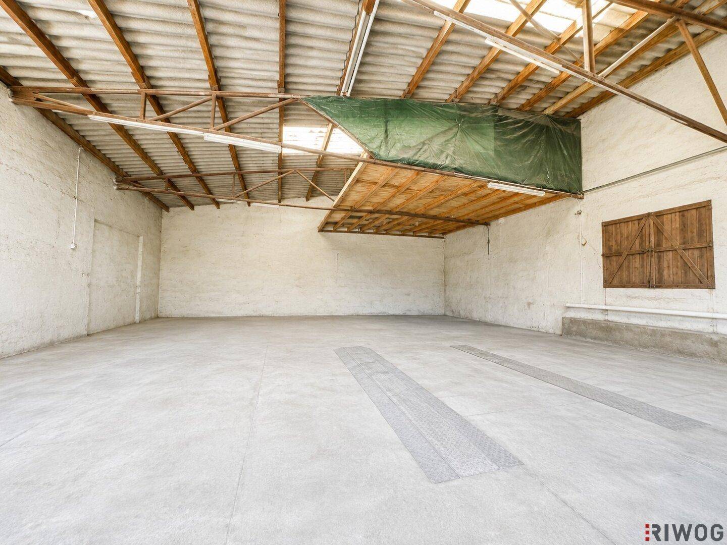 Weitläufiger, leerer Innenraum mit Betonboden, Holzdachkonstruktion und viel Tageslicht.