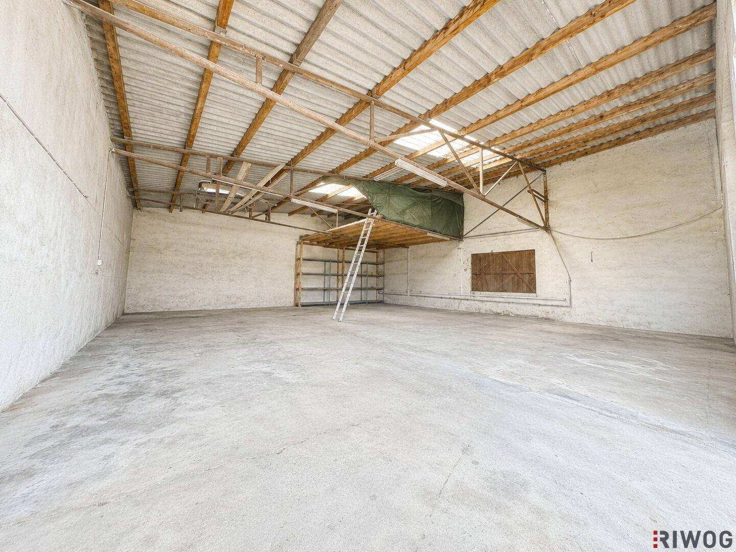 Großer, leerer Innenraum mit hoher Decke, sichtbaren Holzbalken und Betonboden.