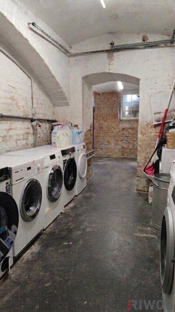 Waschküche im Keller mit mehreren Waschmaschinen, unverputzten Ziegelwänden und einem Fenster im Hintergrund.