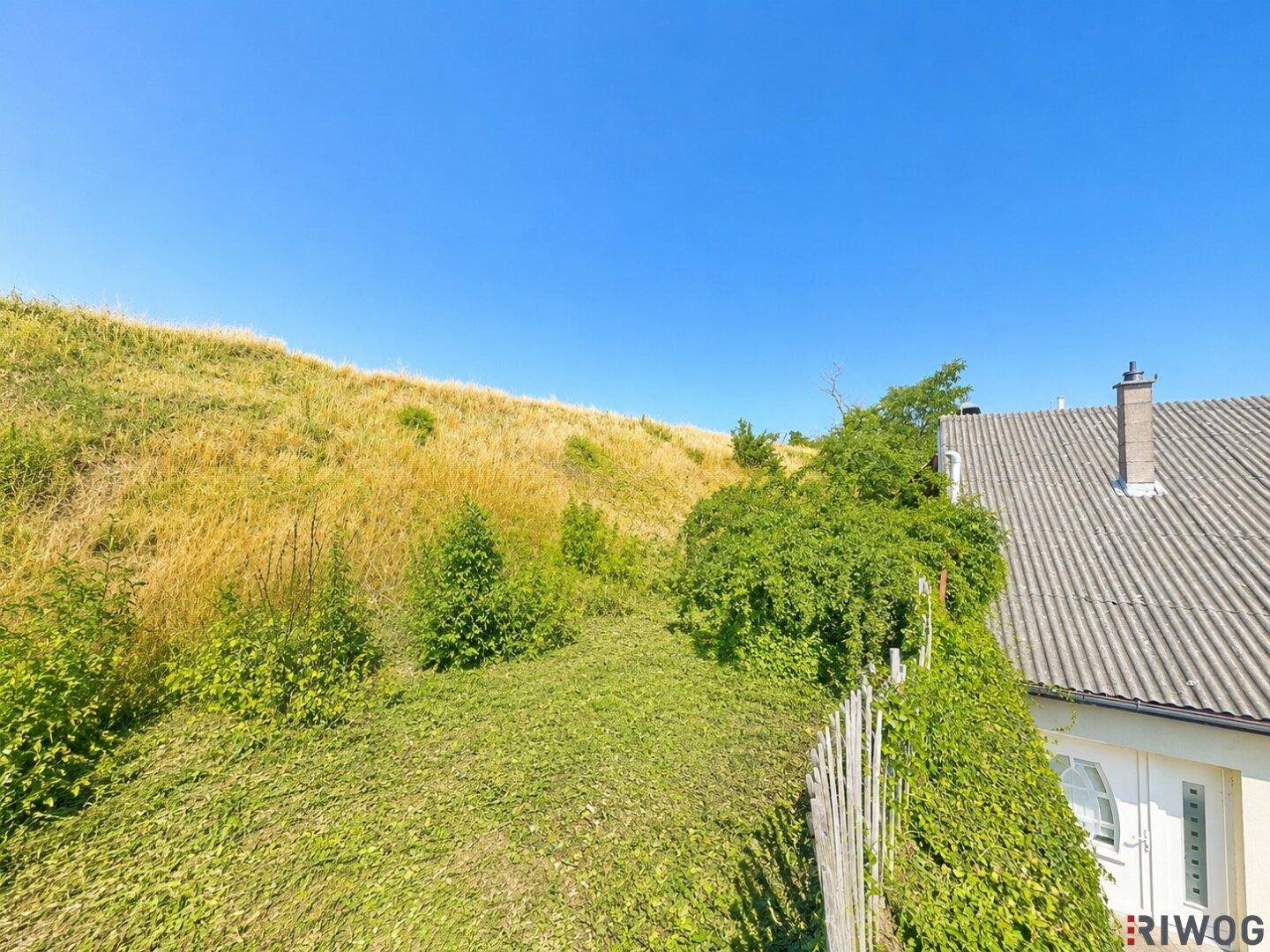 Sanfter grüner Hügel mit üppiger Vegetation und einem Teil eines Hausdaches im Vordergrund.