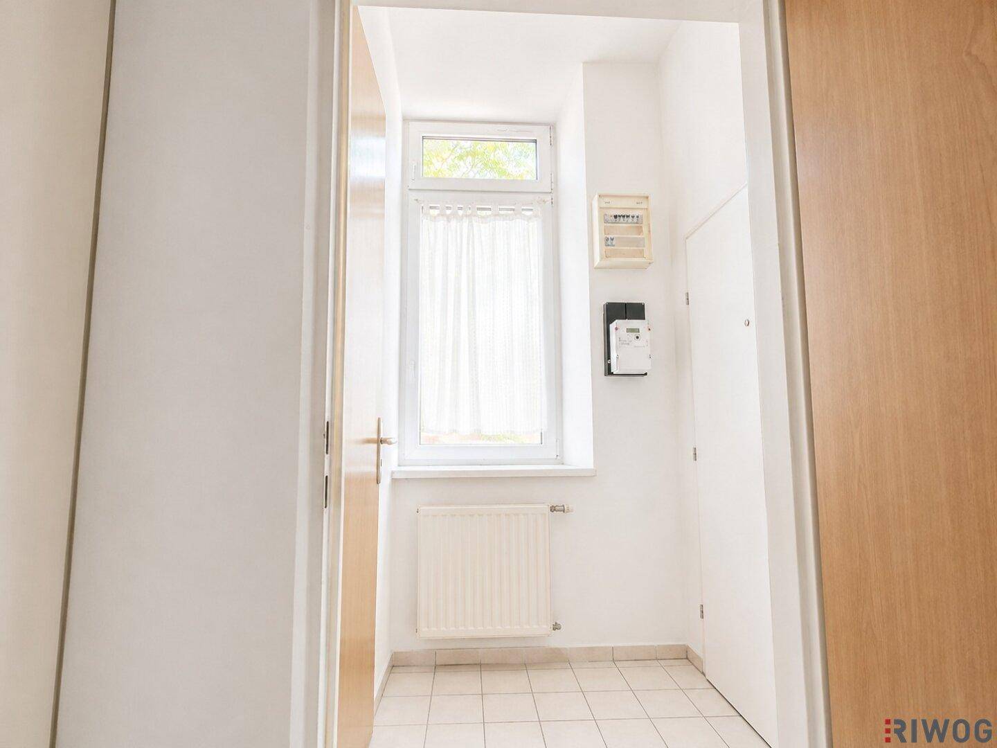 Heller Eingangsbereich mit weiß gefliestem Boden, Fenster und Heizkörper, der zu weiteren Räumen führt.