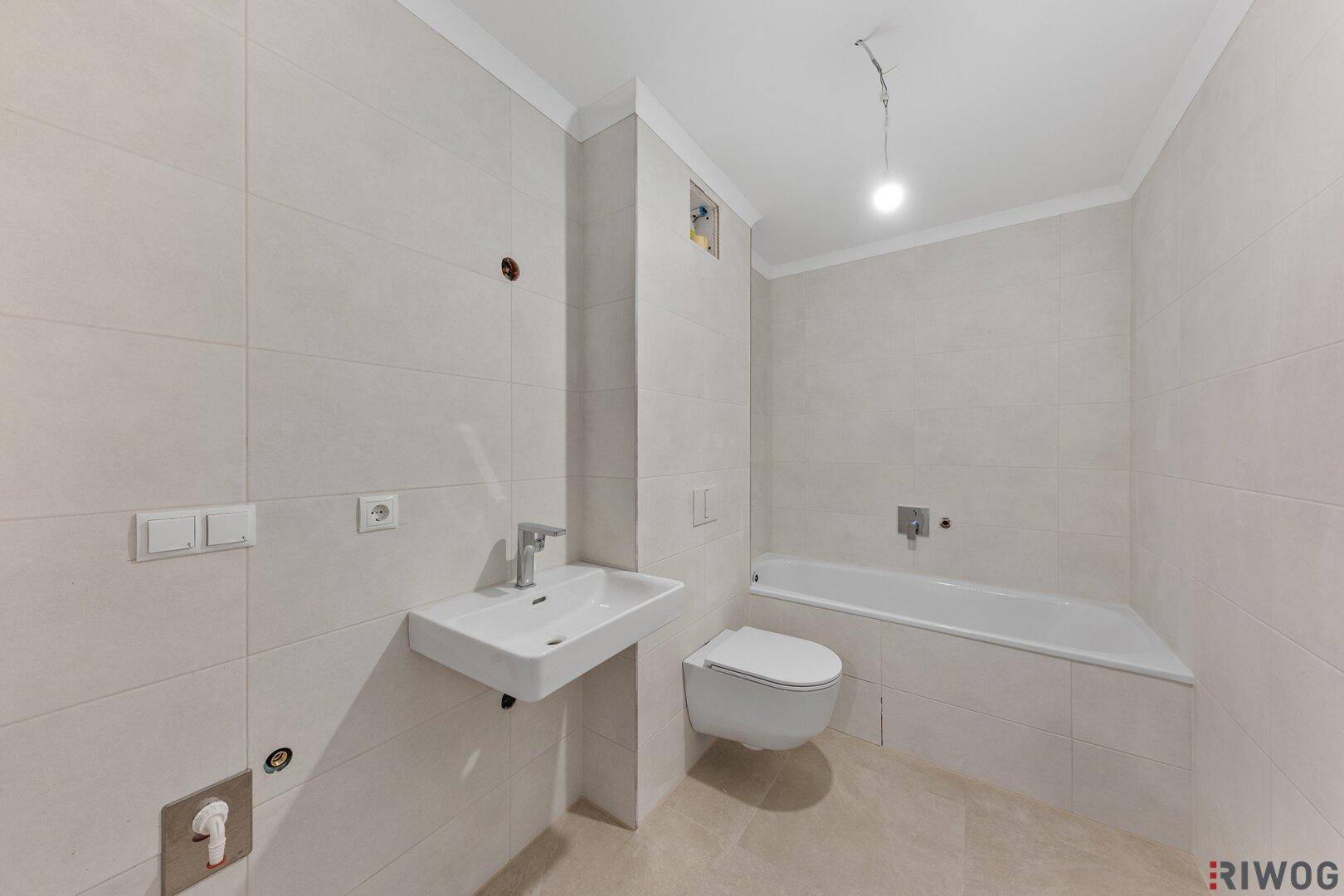 Helles Badezimmer mit weißen Fliesen, wandhängendem WC und Badewanne, noch im Bau.