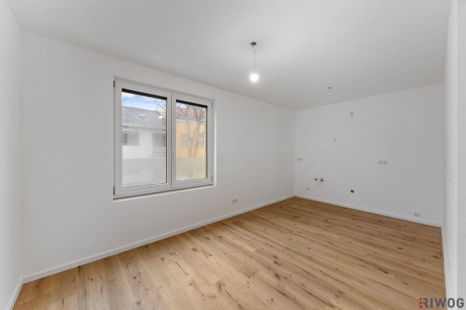 Helles, unmöbliertes Zimmer mit breitem Fenster, Holzboden und weißen Wänden.