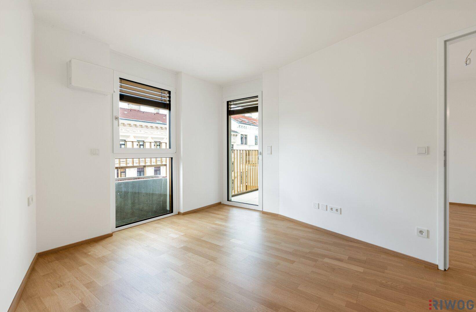 Leeres Schlafzimmer mit Holzboden und zwei großen Fenstern, die viel Licht hereinlassen.