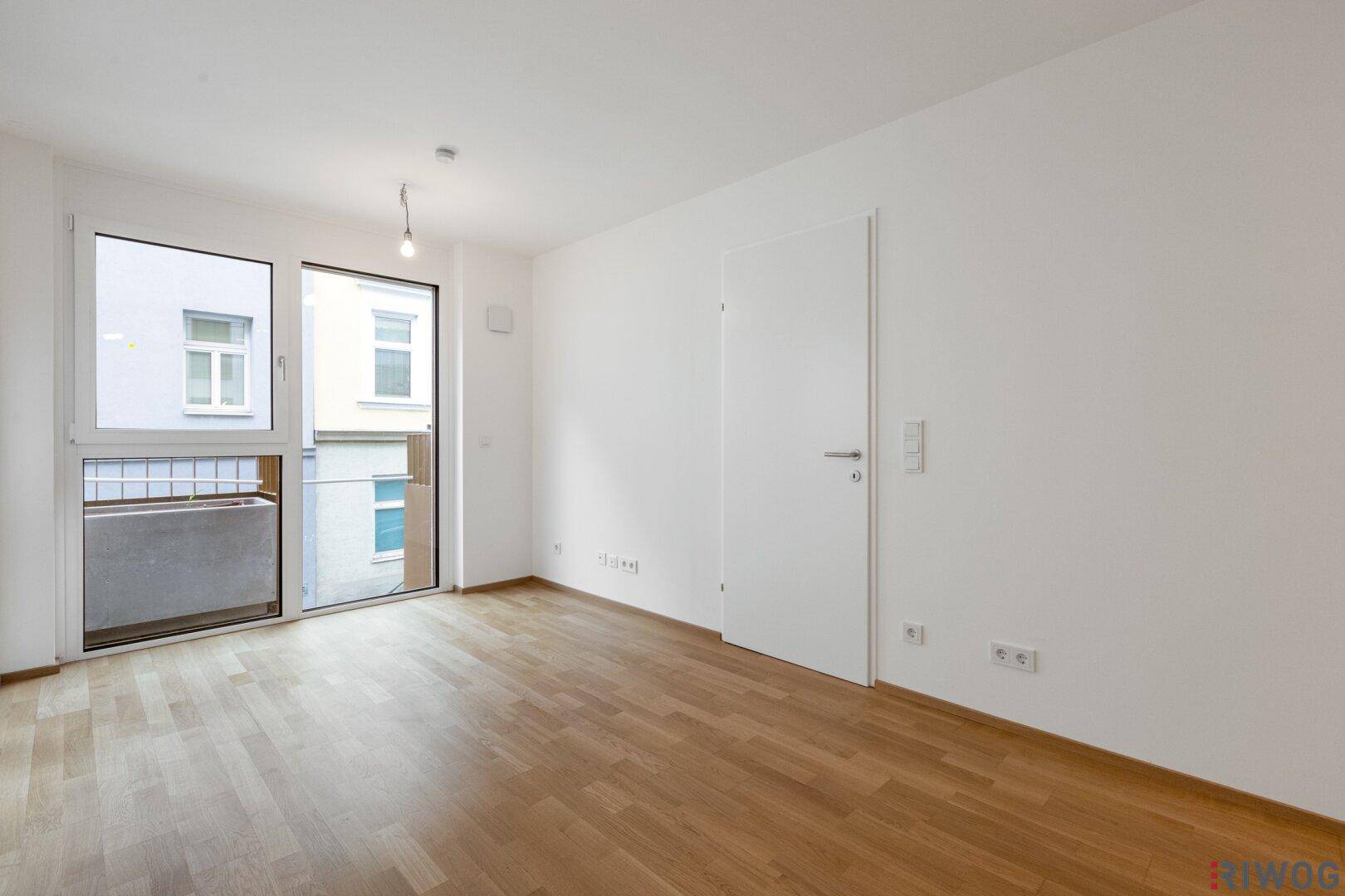 Leeres Schlafzimmer mit hellem Parkettboden und großem Fenster mit Zugang zu einem kleinen Balkon.