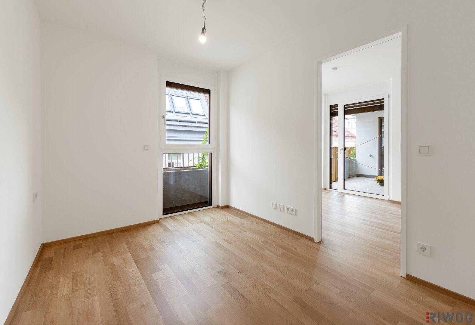 Heller Raum mit Parkettboden und Fenster, der einen Blick in einen weiteren Raum mit Balkonzugang bietet.