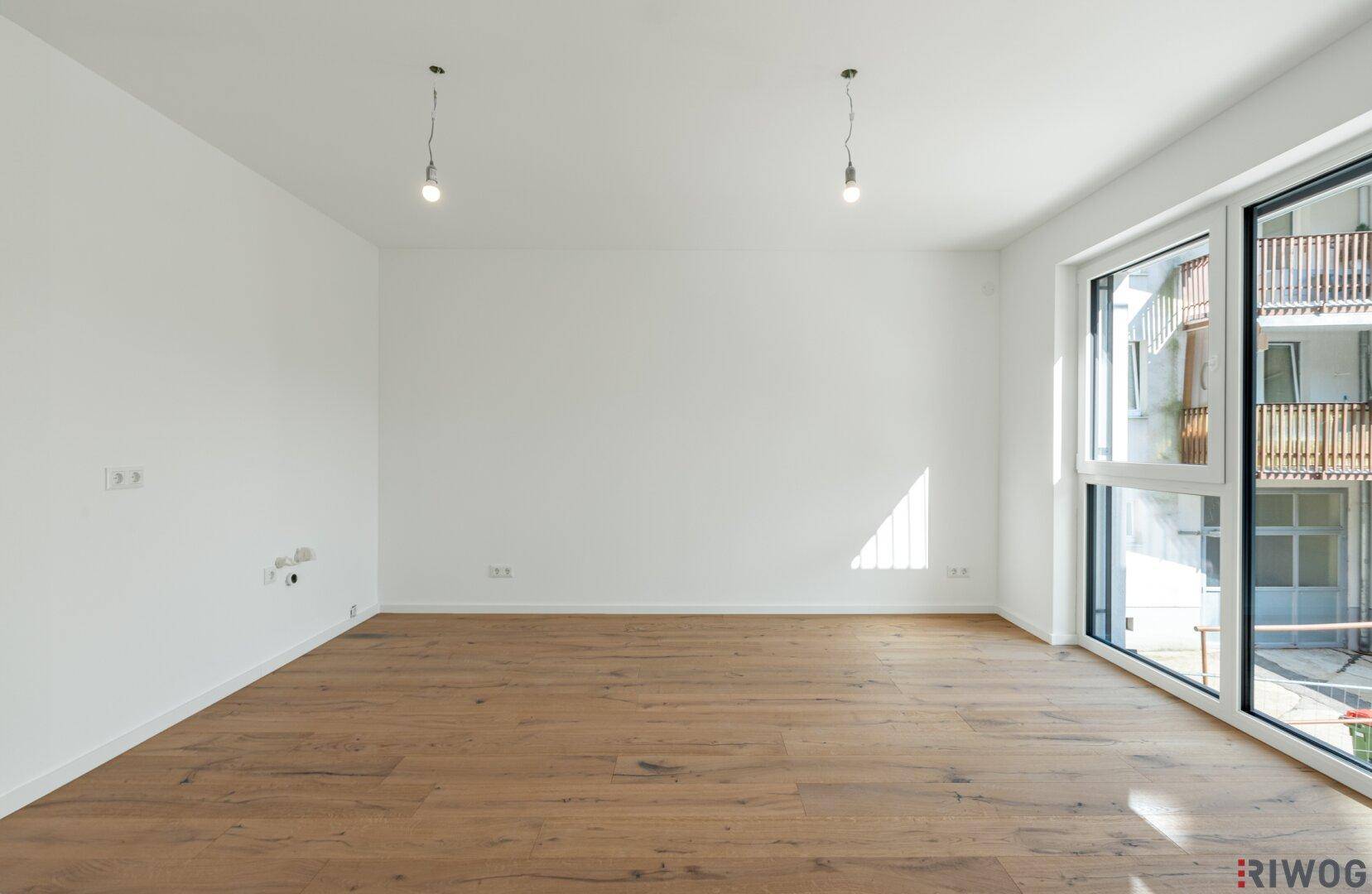 Heller, leerer Raum mit Holzboden und großen Fenstern, die viel Tageslicht hereinlassen.