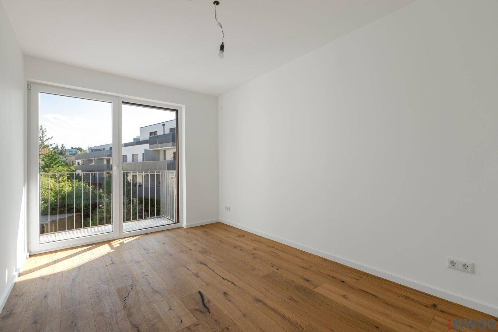 Leeres Schlafzimmer mit Holzboden und großem Fenster, das viel Licht hereinlässt.