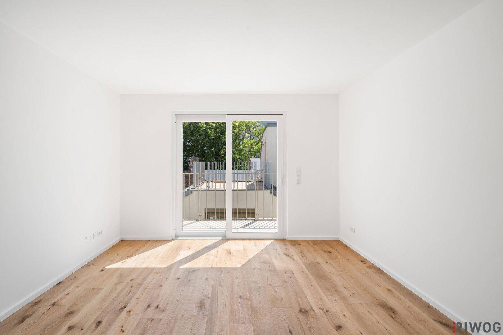 Heller, leerer Raum mit Holzboden und Zugang zu einem Balkon, durch große Schiebetüren.