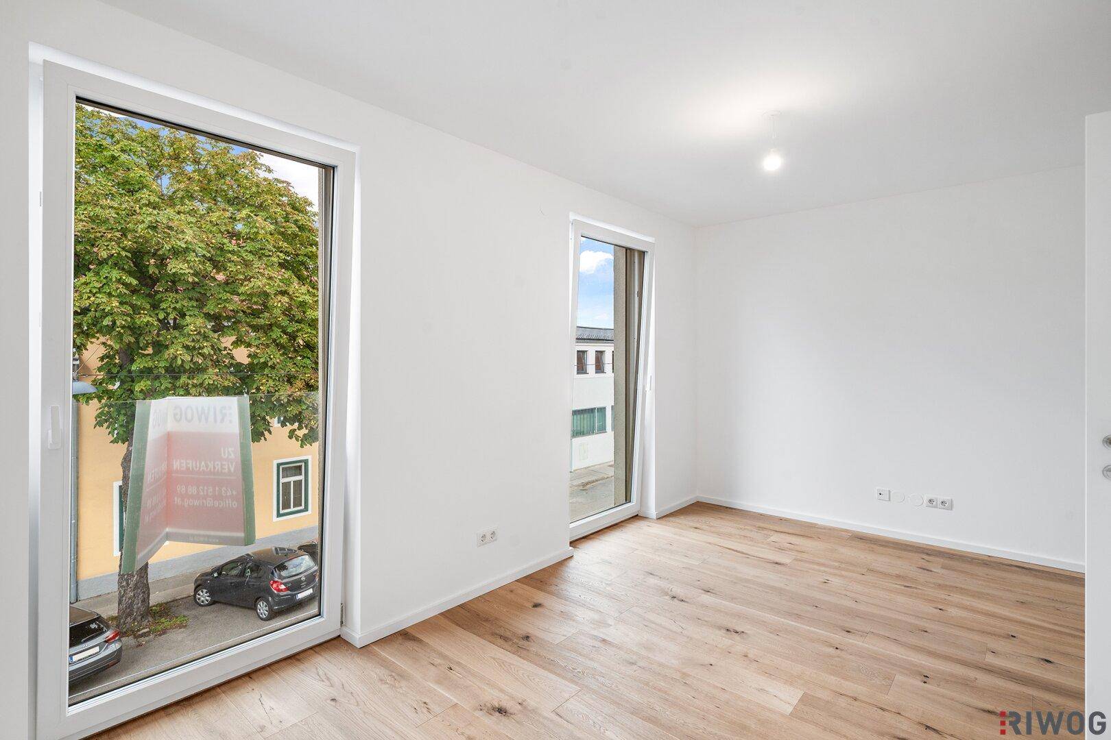 Geräumiges Zimmer mit hellen Wänden und Holzboden, mit Blick auf die Straße und Bäume.