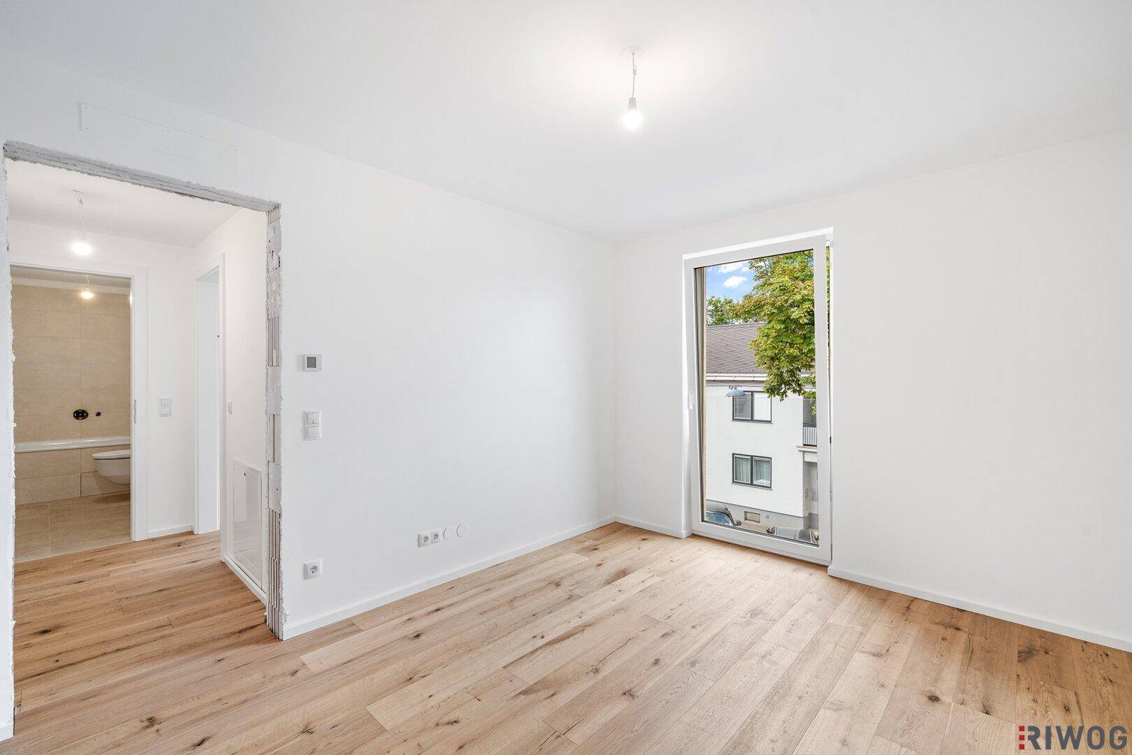 Helles Zimmer mit Holzboden und Zugang zu einem Badezimmer, mit Blick nach draußen.