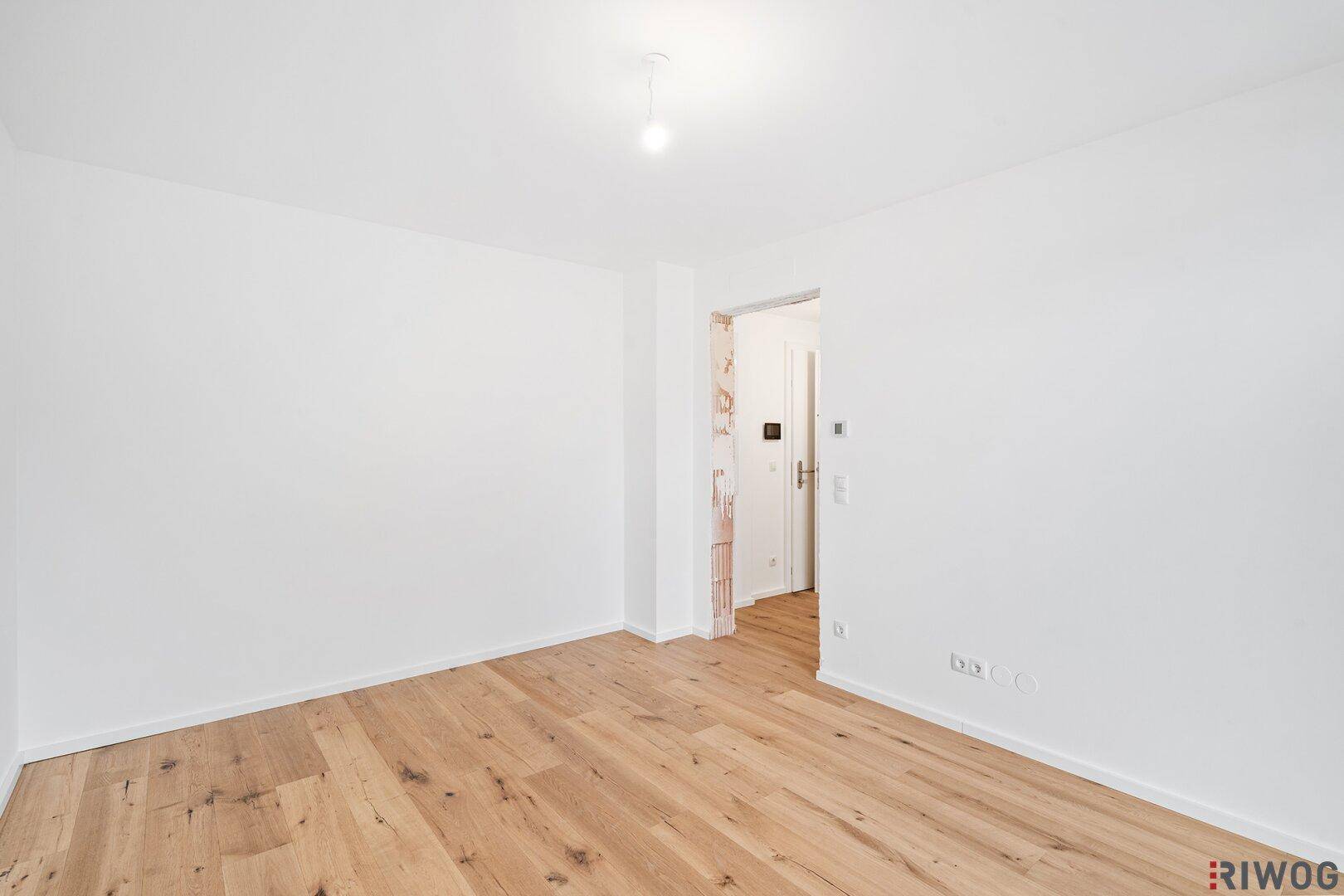 Geräumiges Zimmer mit Holzboden und weiß gestrichenen Wänden, mit Zugang zu einem Flur.