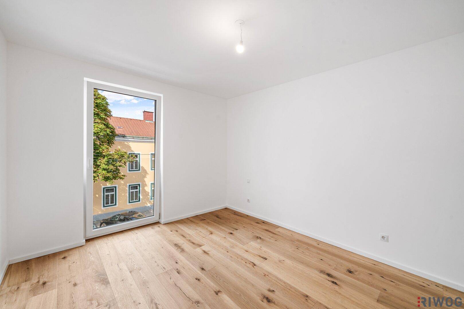 Lichtdurchflutetes Zimmer mit Holzboden und großem Fenster, das einen Blick auf die Umgebung freigibt.
