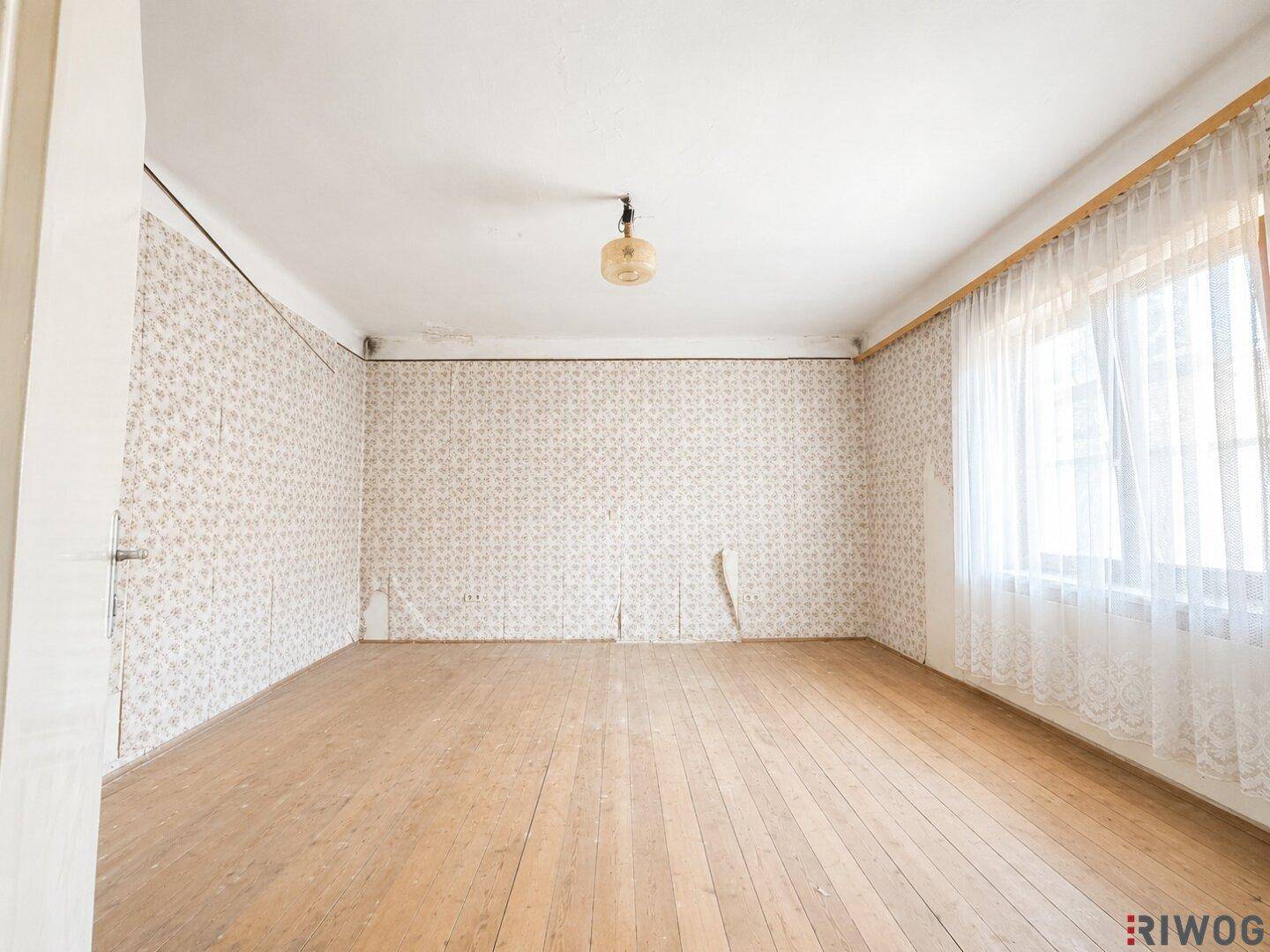 Leeres Zimmer mit gemusterter Tapete, Holzdielenboden und einem großen Fenster mit Vorhängen.
