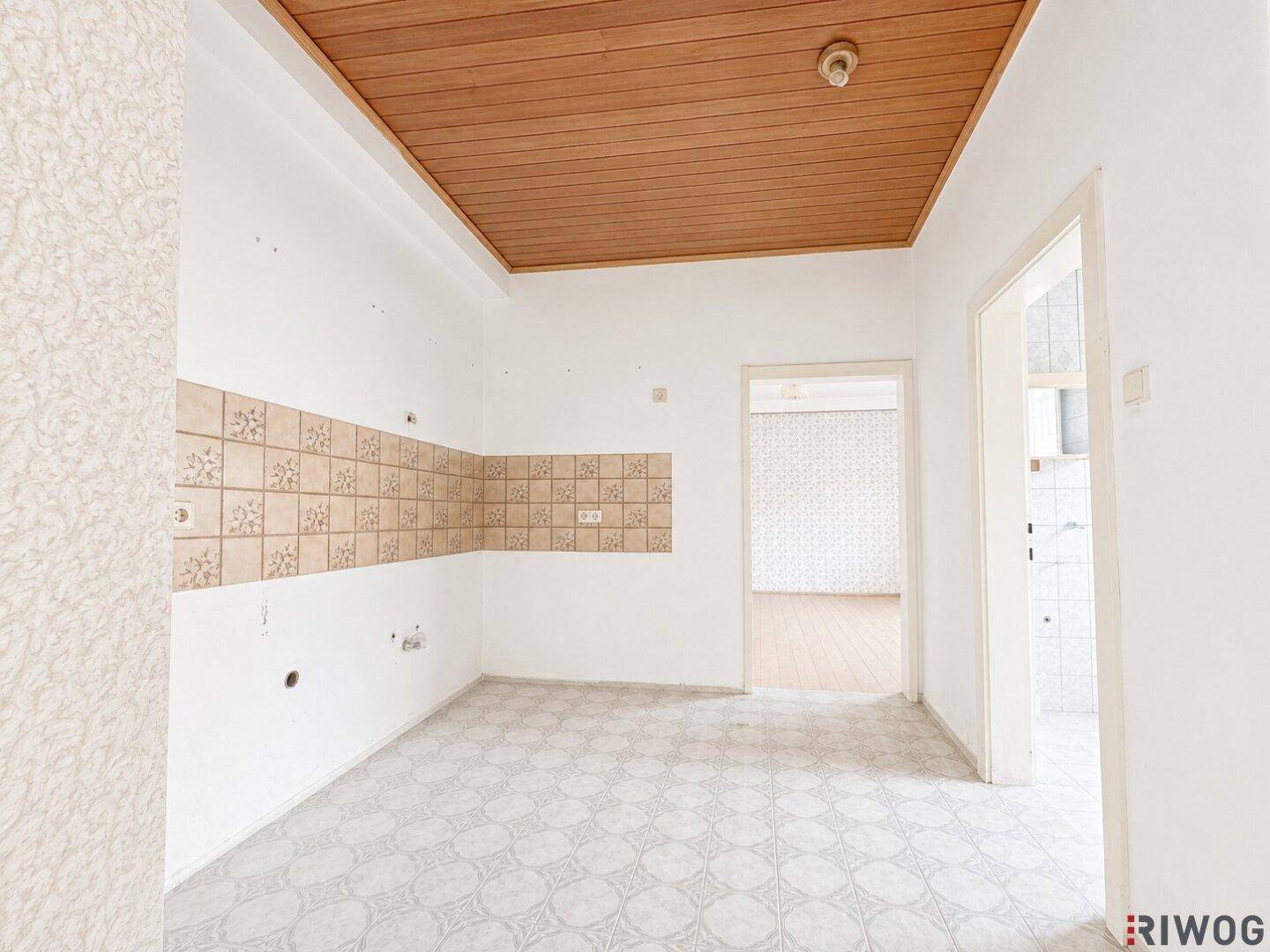 Leere Küche mit Holzdecke, Wandfliesen und Zugang zu weiteren Räumen.