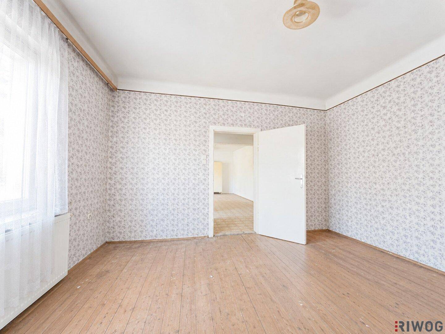 Helles Zimmer mit gemusterter Tapete, Holzdielenboden und einem Fenster mit Vorhängen.