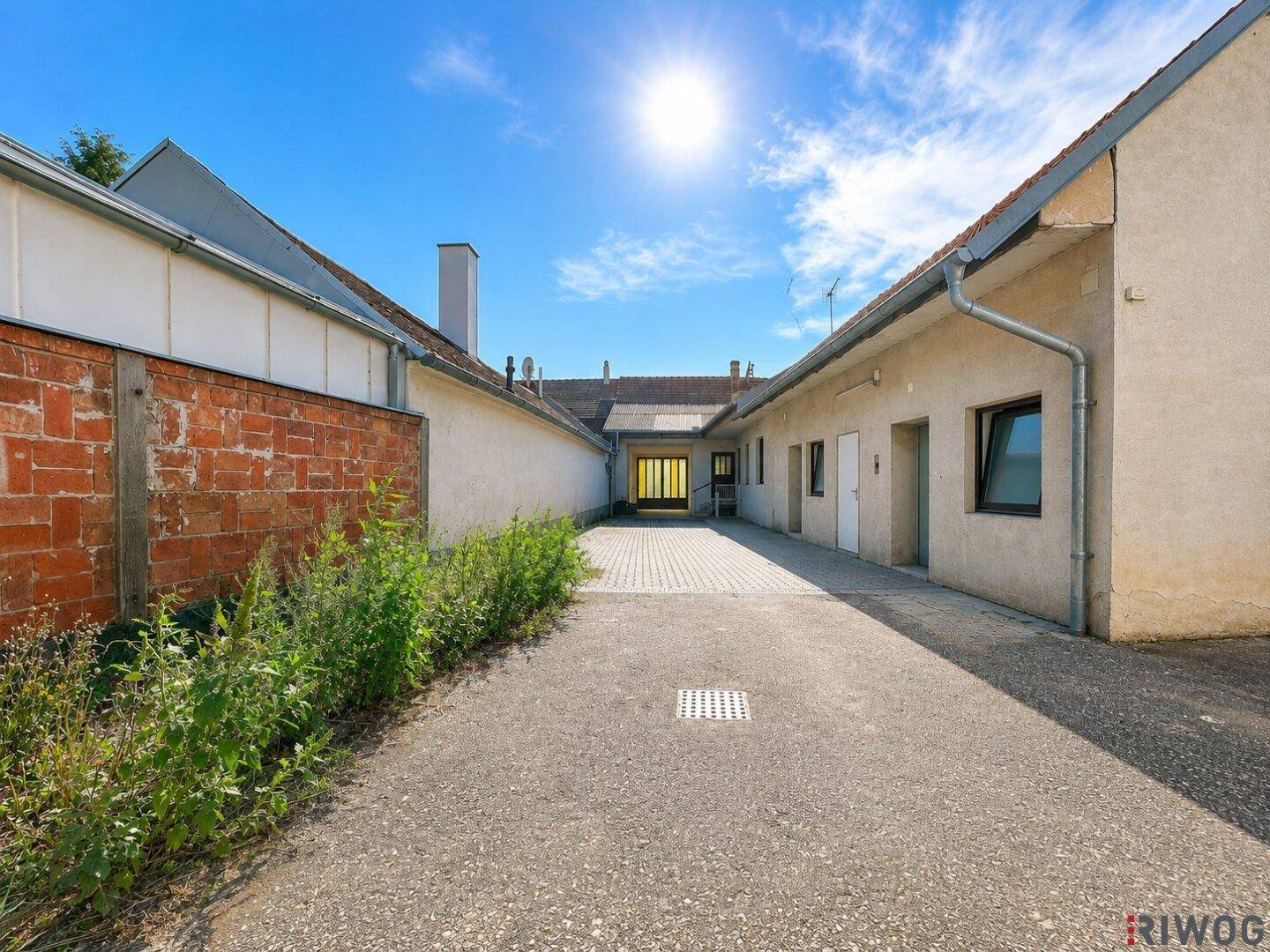 Langer gepflasterter Hofweg zwischen Gebäuden mit einer Ziegelmauer und Büschen.