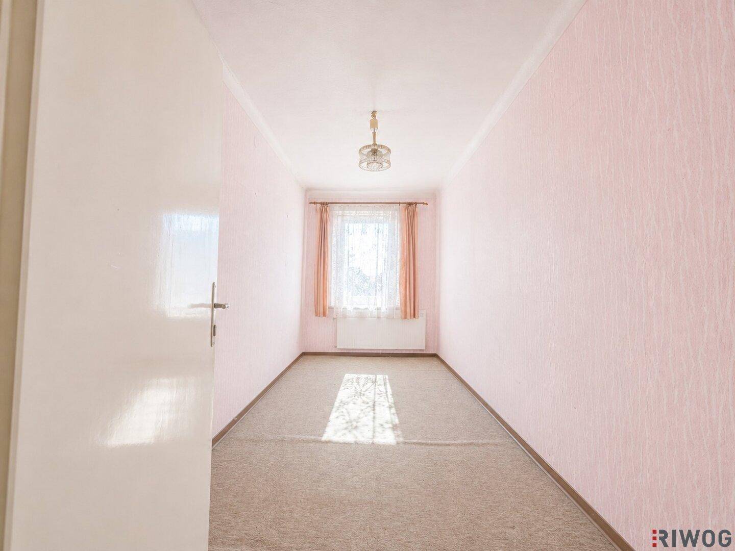 Helles Zimmer mit rosa Tapete, Teppichboden und einem Fenster mit Vorhängen.
