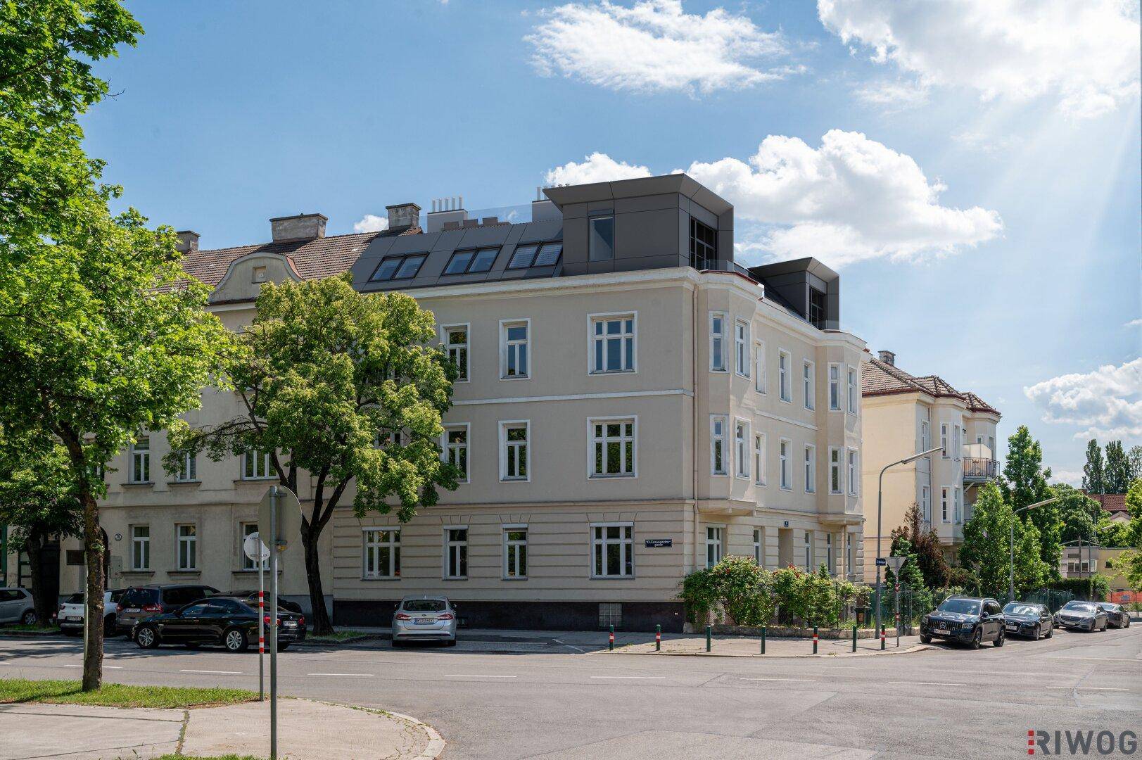 Gebäudeansicht von der Straße mit klassischer Fassade und modernem Dachausbau unter blauem Himmel.