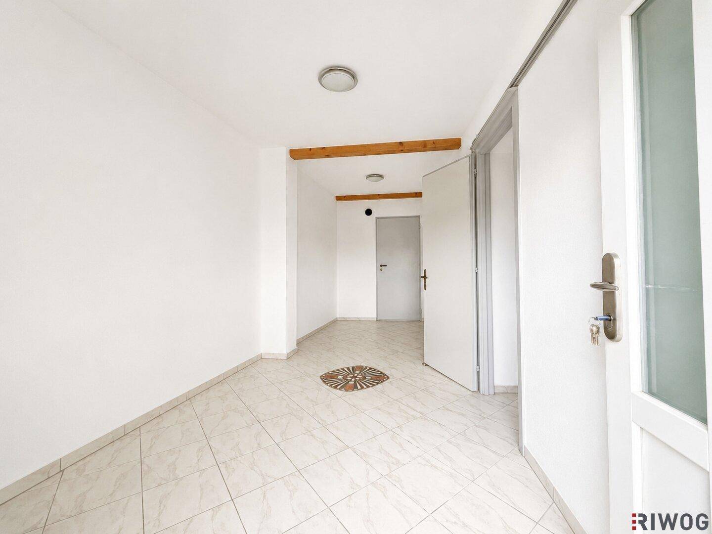 Heller Eingangsbereich mit Fliesenboden, weißen Wänden und sichtbaren Holzbalken an der Decke.